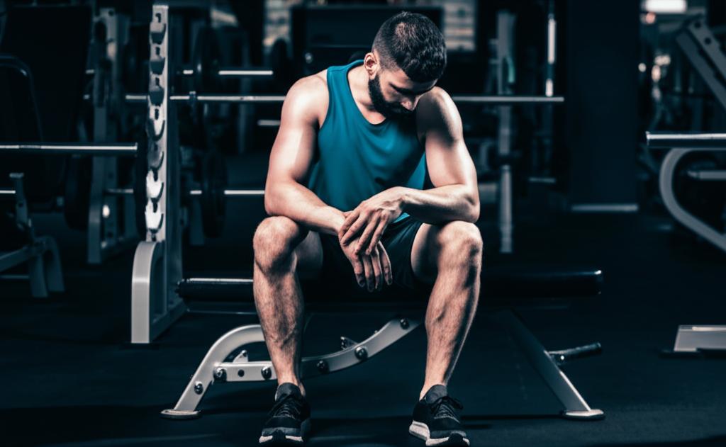 AI prompt: exhausted man sitting on gym bench, looking defeated, low energy, dark lighting