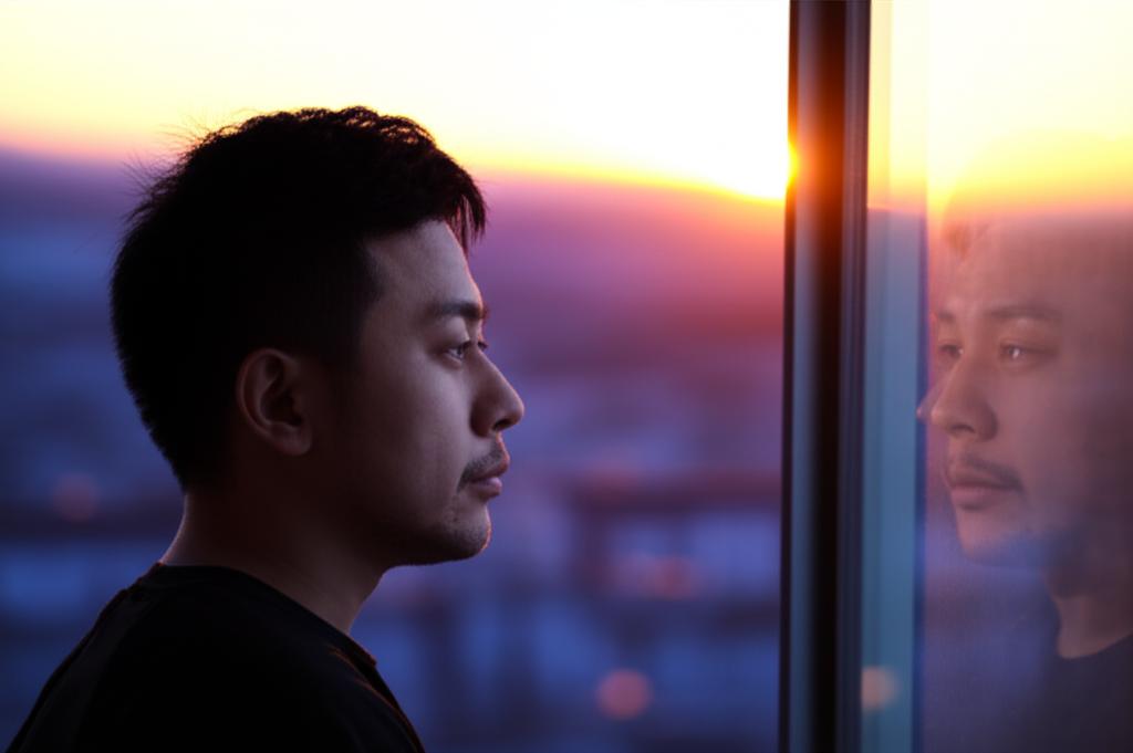 A thoughtful Asian man looking out a window at sunrise, symbolizing reflection on vitality